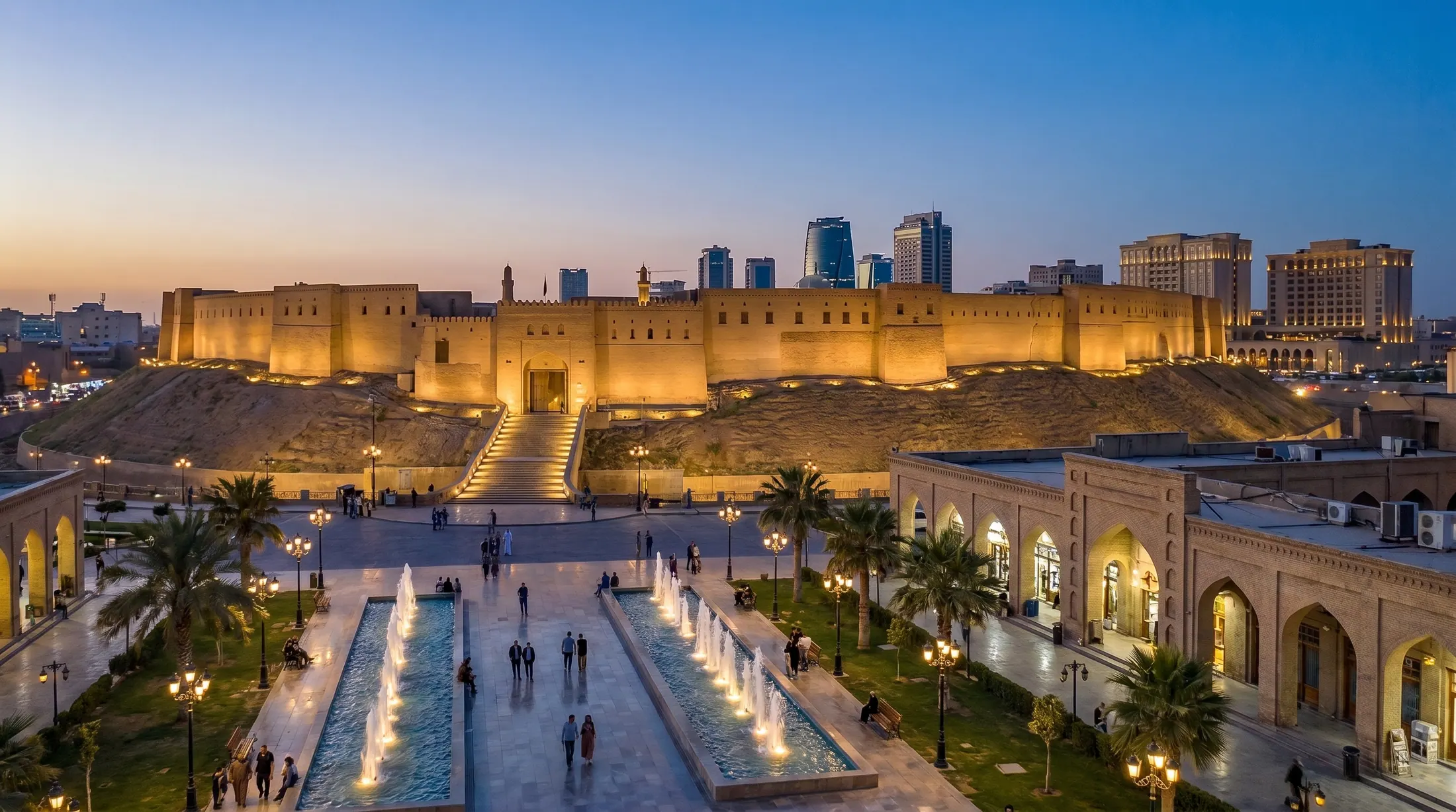 The illuminated Citadel of Erbil at blue hour, the square with water basin and fountains and the bazaar arcades in front, tall buildings and hotels behind